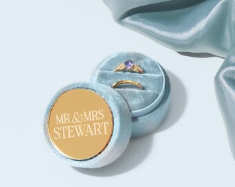 May include: A light blue velvet ring box with a gold lid. The lid has the text "MR & MRS STEWART" engraved on it. The box is open and contains two gold rings.