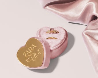 May include: Pink velvet heart-shaped ring box with a gold lid. The lid has the words "ZARA Elliot" engraved on it. The box is open and contains two gold rings.