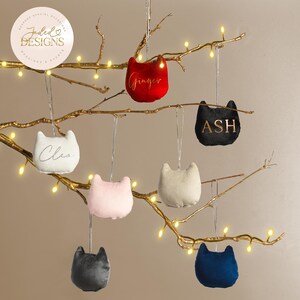 May include: A selection of cat-shaped decorations in shades of red, black, and pink, suspended from a golden branch adorned with fairy lights. Each ornament features a name, like "Ginger" or "Ash". The decorations are crafted from soft fabric.