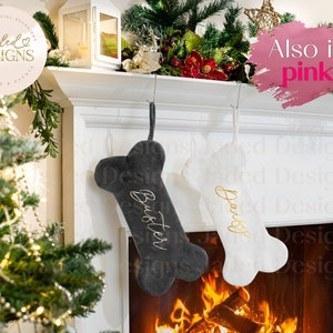 May include: Two personalized dog bone-shaped Christmas stockings hanging on a white fireplace mantel. The stocking on the left is gray with the name "Buster" embroidered in gold. The stocking on the right is white with the name "Daisy" embroidered in gold. The stockings are hanging in front of a fireplace with a roaring fire.