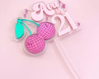 Può includere: Un cake topper in acrilico rosa con il nome "Ava" e il numero "21". Il design include due ciliegie a sfera da discoteca rosa con foglie verdi. Il topper è su un bastoncino trasparente.