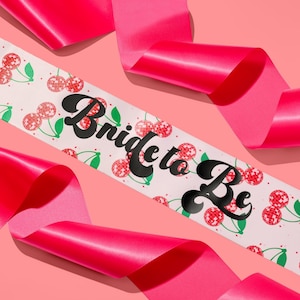 Könnte beinhalten: Weißer Satin-Schärpe mit dem Text "Bride to Be" in schwarzer Schrift. Die Schärpe ist mit roten und weißen Kirschen mit einem Discokugel-Muster verziert. Die Schärpe ist um ein rosa Band gewickelt.