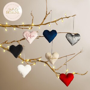 May include: A display of heart-shaped ornaments in shades of pink, navy, black, white, grey, and red, suspended from a gold-painted branch. Some have embroidered names. The ornaments are hung by silver strings.