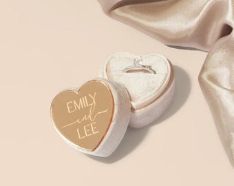 May include: A gold heart-shaped ring box with the inscription "Emily and Lee" on the lid. The box is open and a silver ring is inside.
