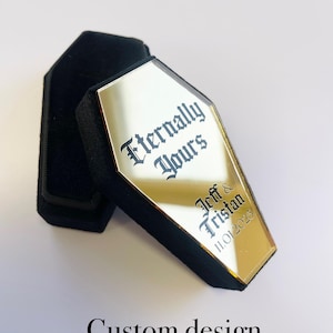 May include: Gold coffin-shaped box with a mirrored surface. The box has the text "Eternally Yours" and "Jeff & Tristan 11.01.2025" engraved on it.