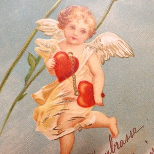 Op de afbeelding: Een vintage Valentijnskaart met een cherubijn met vleugels, die twee rode harten vasthoudt. De engel is gekleed in een witte jurk en heeft een gouden halo. De kaart heeft een blauwe achtergrond met een bloemmotief en de tekst "Je t'embrasse Melanie".