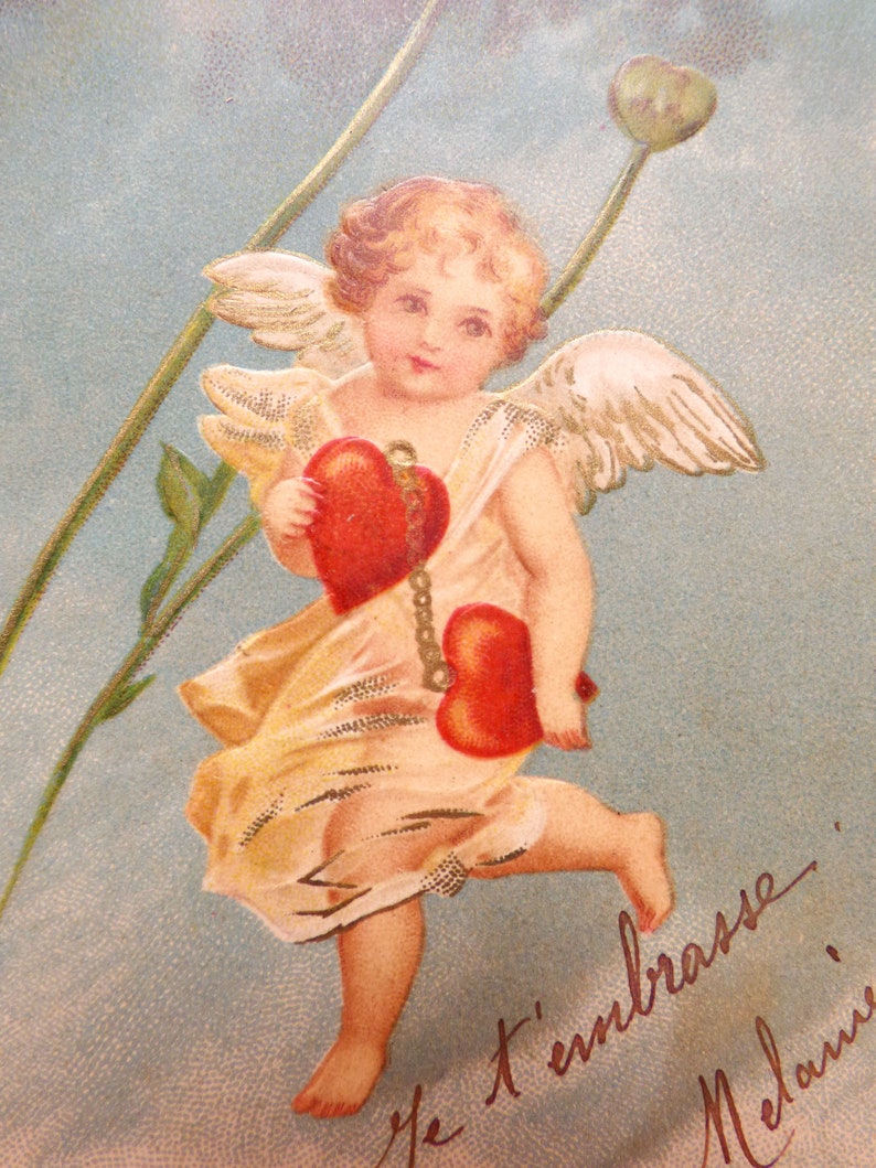 Op de afbeelding: Een vintage Valentijnskaart met een cherubijn met vleugels, die twee rode harten vasthoudt. De engel is gekleed in een witte jurk en heeft een gouden halo. De kaart heeft een blauwe achtergrond met een bloemmotief en de tekst "Je t'embrasse Melanie".