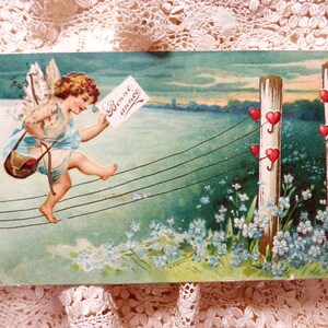 Op de afbeelding: Een vintage ansichtkaart met een cherub met vleugels, die een boog en pijl draagt, en over een draad loopt met een briefje waarop 'Bonne ann&eacute;e' staat. De cherub loopt naar een hek met witte palen waar rode harten aan de draden hangen.