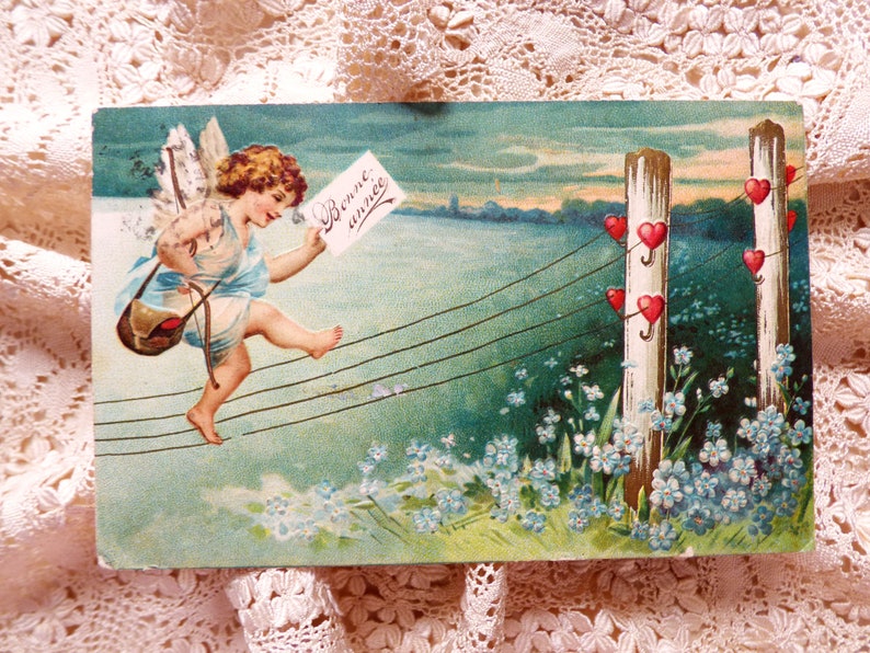 Op de afbeelding: Een vintage ansichtkaart met een cherub met vleugels, die een boog en pijl draagt, en over een draad loopt met een briefje waarop 'Bonne ann&eacute;e' staat. De cherub loopt naar een hek met witte palen waar rode harten aan de draden hangen.