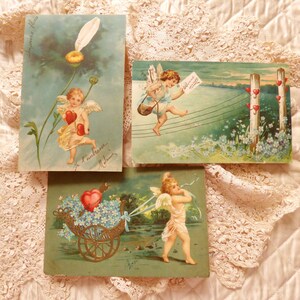 Op de afbeelding: Drie vintage Valentijnsdagpostkaarten met cherubijnen. De eerste kaart toont een cherubijn die twee rode harten vasthoudt en boven een bloem vliegt. De tweede kaart toont een cherubijn die door de lucht vliegt met een brief en een reeks harten. De derde kaart toont een cherubijn die een kar vol bloemen en een hart trekt.