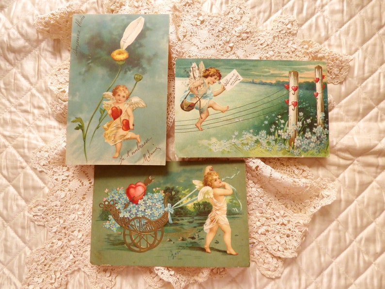 Op de afbeelding: Drie vintage Valentijnsdagpostkaarten met cherubijnen. De eerste kaart toont een cherubijn die twee rode harten vasthoudt en boven een bloem vliegt. De tweede kaart toont een cherubijn die door de lucht vliegt met een brief en een reeks harten. De derde kaart toont een cherubijn die een kar vol bloemen en een hart trekt.