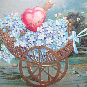 Op de afbeelding: Een vintage Valentijnskaart met een gouden koets gevuld met blauwe vergeet-mij-nietjes en twee rode harten. De koets wordt getrokken door een wit lint en staat tegen een blauwe en groene achtergrond.