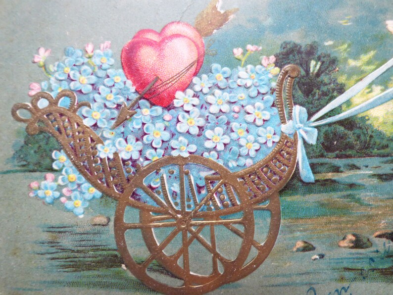 Op de afbeelding: Een vintage Valentijnskaart met een gouden koets gevuld met blauwe vergeet-mij-nietjes en twee rode harten. De koets wordt getrokken door een wit lint en staat tegen een blauwe en groene achtergrond.