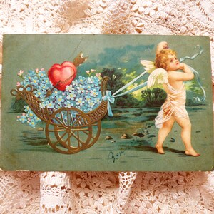 Op de afbeelding: Een vintage Valentijnsdagkaart met een cherub die een kar trekt vol blauwe vergeet-mij-nietjes en een rood hart. De cherub draagt een witte robe en heeft vleugels. De kaart heeft een groene achtergrond en de tekst "Van" staat rechtsonder.