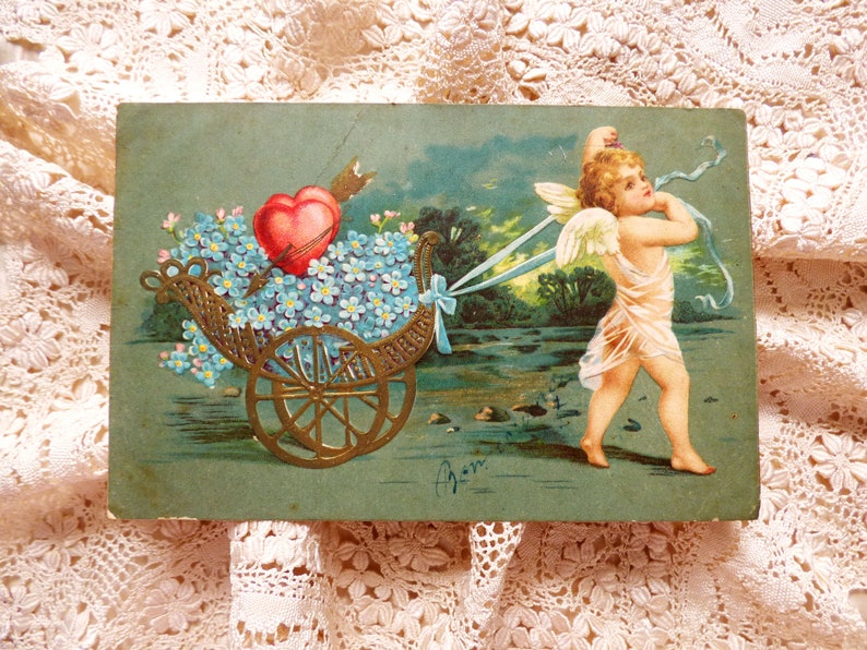 Op de afbeelding: Een vintage Valentijnsdagkaart met een cherub die een kar trekt vol blauwe vergeet-mij-nietjes en een rood hart. De cherub draagt een witte robe en heeft vleugels. De kaart heeft een groene achtergrond en de tekst "Van" staat rechtsonder.