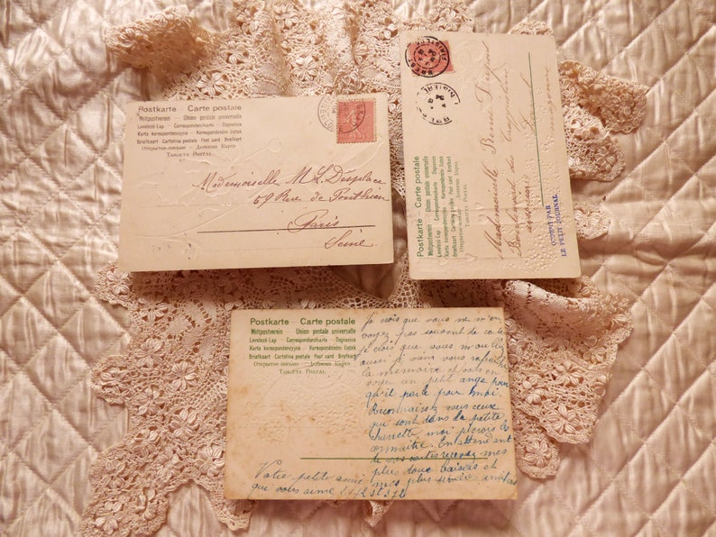 Op de afbeelding: Drie vintage ansichtkaarten met handgeschreven berichten in het Frans. De ansichtkaarten liggen op een kantonderlegger en hebben een cr&egrave;mekleurige achtergrond. De tekst op de ansichtkaarten luidt "Postkarte Carte postale" en "Mademoiselle M.R. Despedes 67 Rue de Freithin Paris".