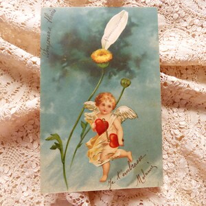 Op de afbeelding: Een vintage ansichtkaart met een cherub met vleugels die twee rode harten vasthoudt. De cherub vliegt boven een blauwe lucht met witte bloemen. De tekst op de ansichtkaart luidt "Je t'embrasse Melanie".