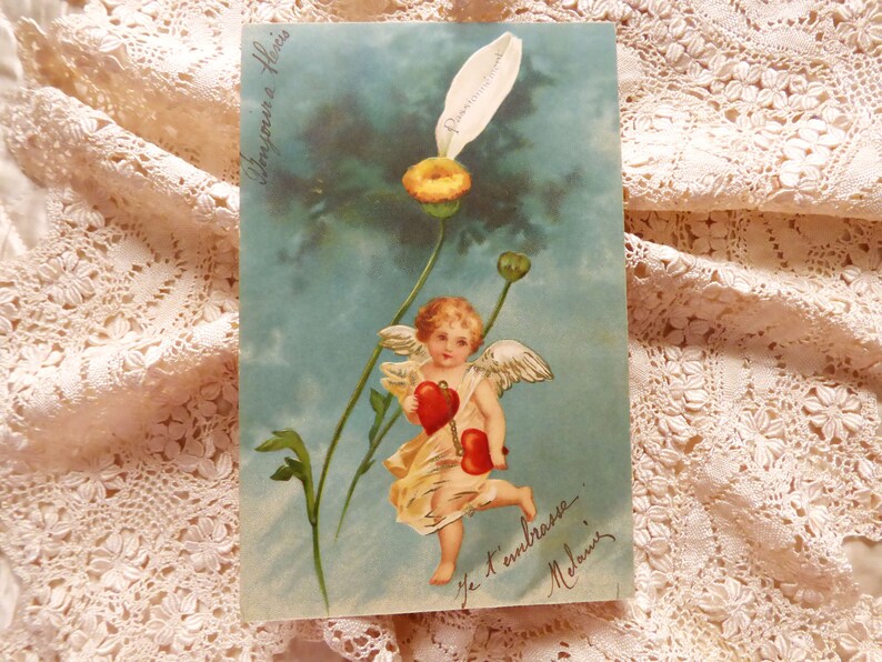Op de afbeelding: Een vintage ansichtkaart met een cherub met vleugels die twee rode harten vasthoudt. De cherub vliegt boven een blauwe lucht met witte bloemen. De tekst op de ansichtkaart luidt "Je t'embrasse Melanie".