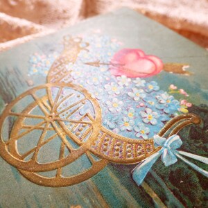 Op de afbeelding: Een vintage Valentijnskaart met een gouden koets gevuld met blauwe vergeet-mij-nietjes en een roze hart met een pijl erdoorheen.