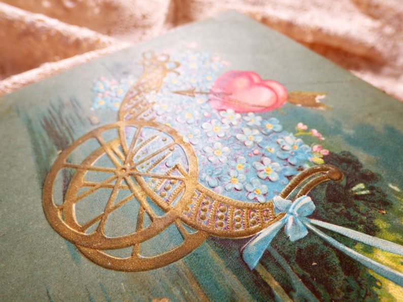 Op de afbeelding: Een vintage Valentijnskaart met een gouden koets gevuld met blauwe vergeet-mij-nietjes en een roze hart met een pijl erdoorheen.