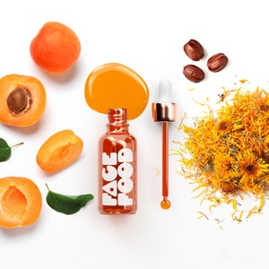 Może przedstawiać: Przezroczysta szklana butelka z kroplomierzem wypełniona pomarańczowym płynem. Butelka jest oznaczona etykietą "FACE FOOD". Na zdjęciu znajdują się również suszone kwiaty pomarańczy, połówki moreli i suszone pestki moreli.