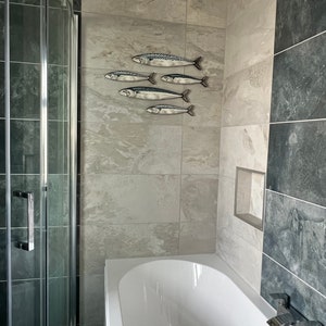 May include: A bathroom scene featuring a white bathtub, a glass shower enclosure, and a wall-mounted decorative fish sculpture. The fish are silver with black and blue details, arranged in a school-like formation on the tiled wall.