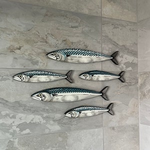 May include: Six decorative fish sculptures with a mackerel design. The fish are painted in shades of silver, blue, and black, with detailed scales and fins. They are mounted on a gray tiled wall, creating a coastal or nautical theme.