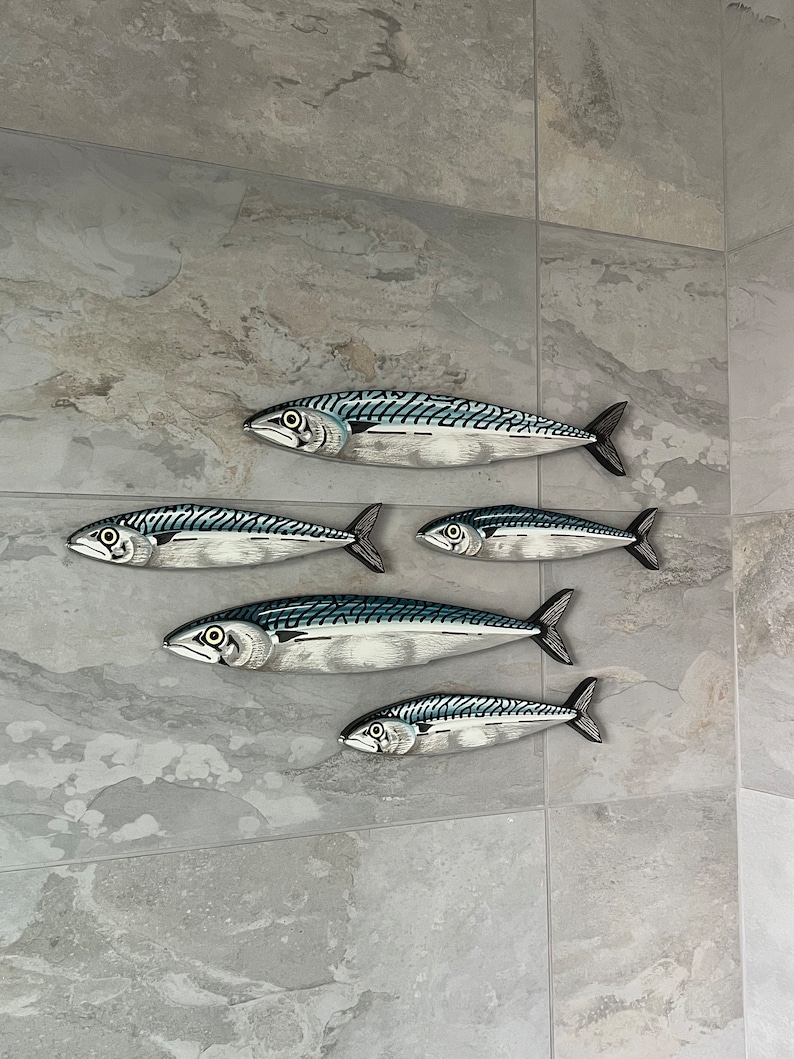 May include: Six decorative fish sculptures with a mackerel design. The fish are painted in shades of silver, blue, and black, with detailed scales and fins. They are mounted on a gray tiled wall, creating a coastal or nautical theme.