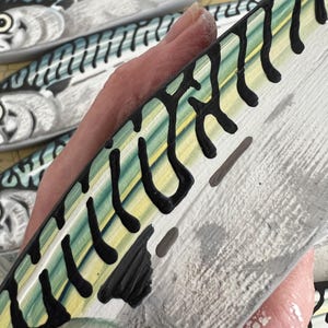 May include: Close-up of a hand holding a painted wooden fish. The fish has a white base with black, yellow, and green markings. Other similar fish are visible in the background, suggesting a nautical theme.