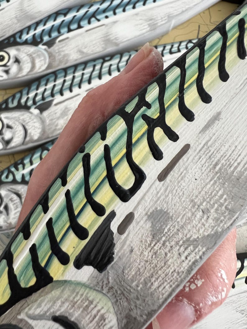 May include: Close-up of a hand holding a painted wooden fish. The fish has a white base with black, yellow, and green markings. Other similar fish are visible in the background, suggesting a nautical theme.