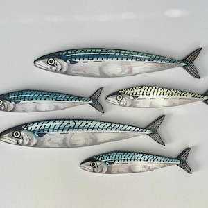 May include: Five painted wooden mackerel fish with detailed scales and fins. The fish are in shades of silver, blue, and green, with black accents. The fish vary in size and are arranged on a white background.