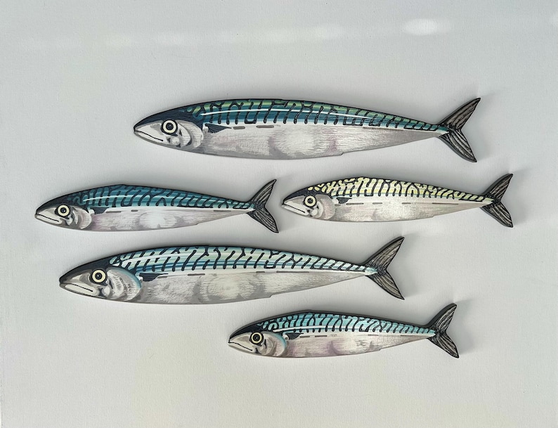 May include: Five painted wooden mackerel fish with detailed scales and fins. The fish are in shades of silver, blue, and green, with black accents. The fish vary in size and are arranged on a white background.