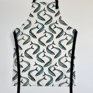 Puede incluir: Delantal blanco con un patrón repetido de peces estilizados en tonos azules, verdes y grises. El delantal tiene correas y lazos negros. Ideal para cocinar o manualidades. Un artículo de cocina o de manualidades.