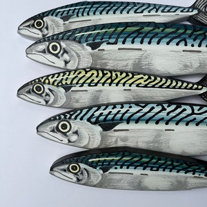 May include: Five wooden mackerel fish, painted in shades of gray, blue, and black, are arranged in a diagonal row. Each fish has detailed markings and a realistic appearance, with visible eyes and fins. The background is plain white.