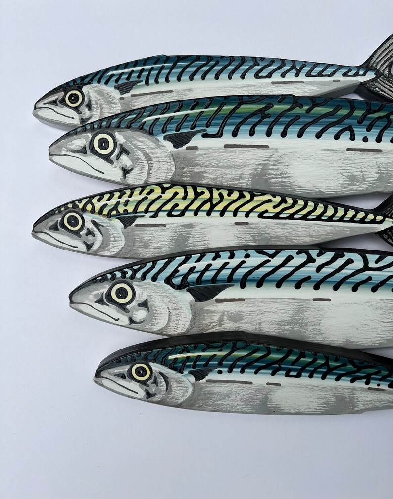 May include: Five wooden mackerel fish, painted in shades of gray, blue, and black, are arranged in a diagonal row. Each fish has detailed markings and a realistic appearance, with visible eyes and fins. The background is plain white.
