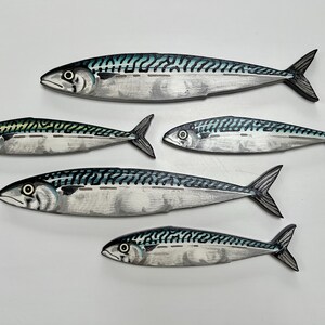 May include: Five wooden mackerel fish sculptures. Each fish has a silver body with black and teal markings. The fish are arranged on a white surface, with the largest fish at the top and the smallest at the bottom.