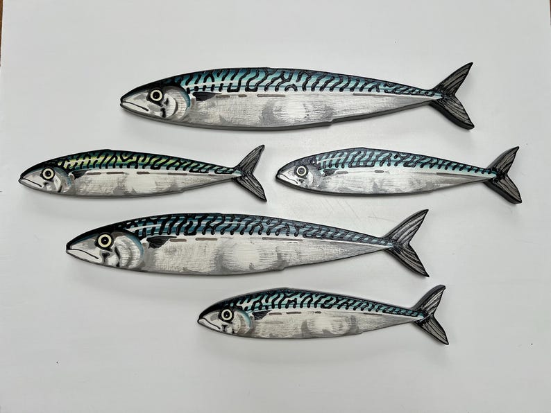 May include: Five wooden mackerel fish sculptures. Each fish has a silver body with black and teal markings. The fish are arranged on a white surface, with the largest fish at the top and the smallest at the bottom.