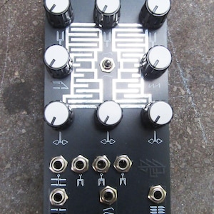 Può includere: Dispositivo elettronico nero con manopole bianche e jack argentati. Il dispositivo ha la scritta "VERATRUM" in alto e "REVERSELANDFILL" in basso. Il dispositivo ha una serie di linee e simboli bianchi.