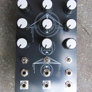 Può includere: Un dispositivo elettronico nero con manopole bianche e jack argentati. Il dispositivo presenta il testo "TRIPLE FUNCTION GENERATOR" e "REVERSELANDFILL" stampato. Il design include linee e cerchi geometrici bianchi.