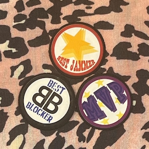 Op de afbeelding: Drie ronde patches met tekst en afbeeldingen. Eén patch heeft een gele ster en de woorden "BEST JAMMER" in het rood. Een andere heeft "BEST BLOCKER" en de letters "BB". De derde patch heeft "MVP" en sterren.