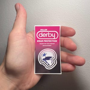 Könnte beinhalten: Ein rosa-schwarzer Aufkleber mit dem Wort "derby" in Weiß und den Worten "WEAR PROTECTION" darunter. Eine Grafik eines Roller-Derby-Helms mit einem Stern befindet sich unten. Der Aufkleber wird in einer Hand gehalten.