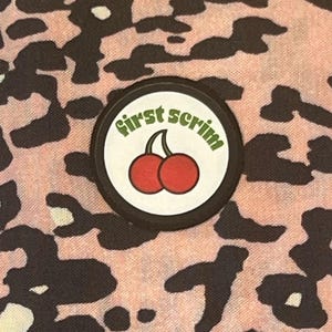 Könnte beinhalten: Nahaufnahme eines Stoffes mit rosa-schwarzem Leopardenmuster. Ein kreisförmiger Aufnäher mit den grünen Worten "first scrim" umgibt ein Paar roter Kirschen.
