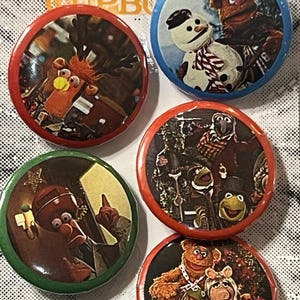 Op de afbeelding: Set van vijf ronde buttons met Muppets-personages. Elke button heeft een andere afbeelding, waaronder Kermit de Kikker, Miss Piggy en Fozzie Beer. De buttons hebben rode, groene en blauwe randen. De tekst "THE PRINTING HUB" is zichtbaar.
