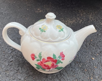 May include: White ceramic teapot with a floral design featuring red and pink peonies, yellow flowers, and green leaves. The teapot has a gold trim, a curved handle, and a lid with a small knob. The spout has a small chip.