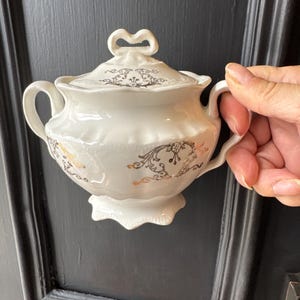 May include: White porcelain sugar bowl with lid. The bowl features a heart-shaped handle on the lid and two side handles. It is decorated with gold and black floral patterns. The sugar bowl sits on a decorative base.