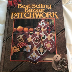 May include: A book titled "Best-Selling Bazaar Patchwork" with a brown wooden background. The cover features a patchwork quilt with star and geometric patterns, pumpkins, and flowers. A small doll is in the lower right corner.