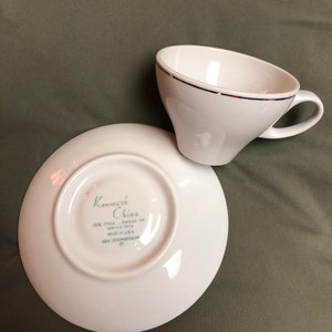 May include: White teacup and saucer set. The teacup has a black trim and a unique angled design. The saucer has the text "Kenmark China" and other text. Made in the USA.