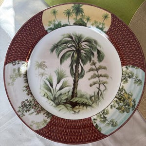 May include: A decorative plate with a tropical palm tree design. The center of the plate features a detailed illustration of a palm tree and other plants. The outer rim has a woven pattern in a reddish-brown color, with sections of tropical scenery.