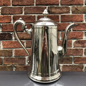 May include: A tall, silver-colored coffee pot with a decorative handle and spout. The pot has a lid with a finial and is set against a brick wall background. The pot is made of a shiny metal.