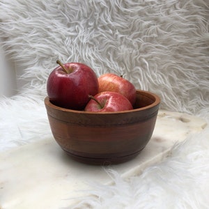 Op de afbeelding: Een houten kom gevuld met drie rode appels. De kom heeft een donkerbruin houtnerfpatroon en staat op een wit, pluche oppervlak.